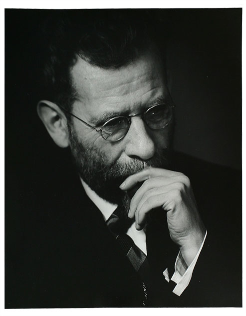 Portrait photograph of an elderly man with a beard and glasses deep in thought touching his fingers to his mouth.