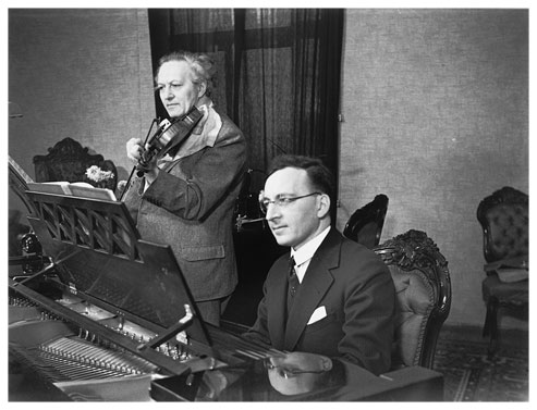 Black-and-white photograph of two men performing in an elegant setting. The younger man wearing glasses and a suit is seated at an open grand piano while the older musician, with his striking white curly hair, is standing next to him playing the violin.