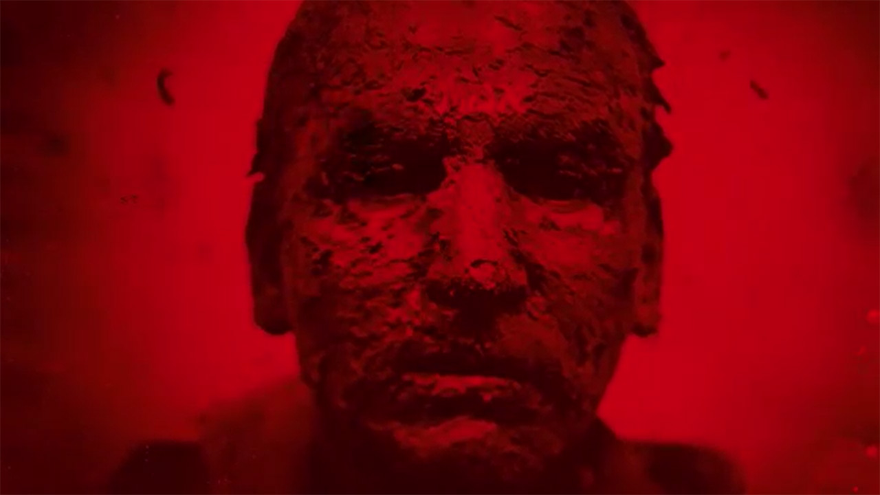 Image dipped in red: a male face smeared with clay.