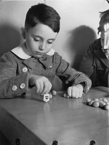 Schwarz-Weiß-Fotografie, die einen Jungen mit Dreidel an einem Tisch zeigt. Am Bildrand ist ein zweiter Junge zu sehen.