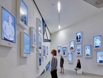 Visitors in an exhibition room with screens scattered along the walls showing videos of various people
