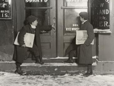 Black and white photograph of two children standing in front of a door with newspapers under their arms.