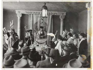 Schwarz-weiß Fotografie einer Synagoge, in der Mitte ein Rabbi, um ihn herum Männer mit Judaica 