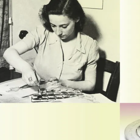 A woman is sitting at her desk covered with colours and brushes. She is painting.