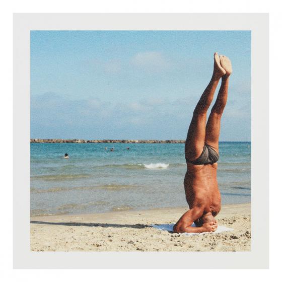 Vintage photograph of the back of a muscular, tan, older man doing a handstand by the shore of a beach