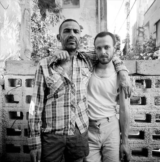 Black and white portrait photo of two men with beards standing in front a cinderblock wall with their arms around each other 