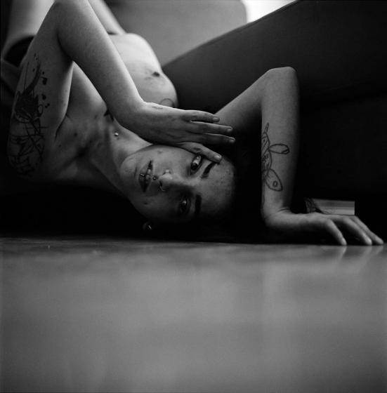 Black and white photograph of a topless woman with multiple piercings and tattoos laying on the floor with her arms framing her face