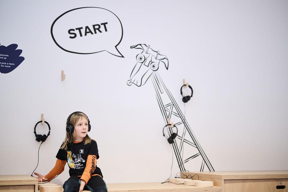 Kind mit Kopfhörern sitzt auf einer Holzkiste. Auf der Rückwand sieht man eine gezeichnete Giraffe mit einer Sprechblase, in der "Start" steht.