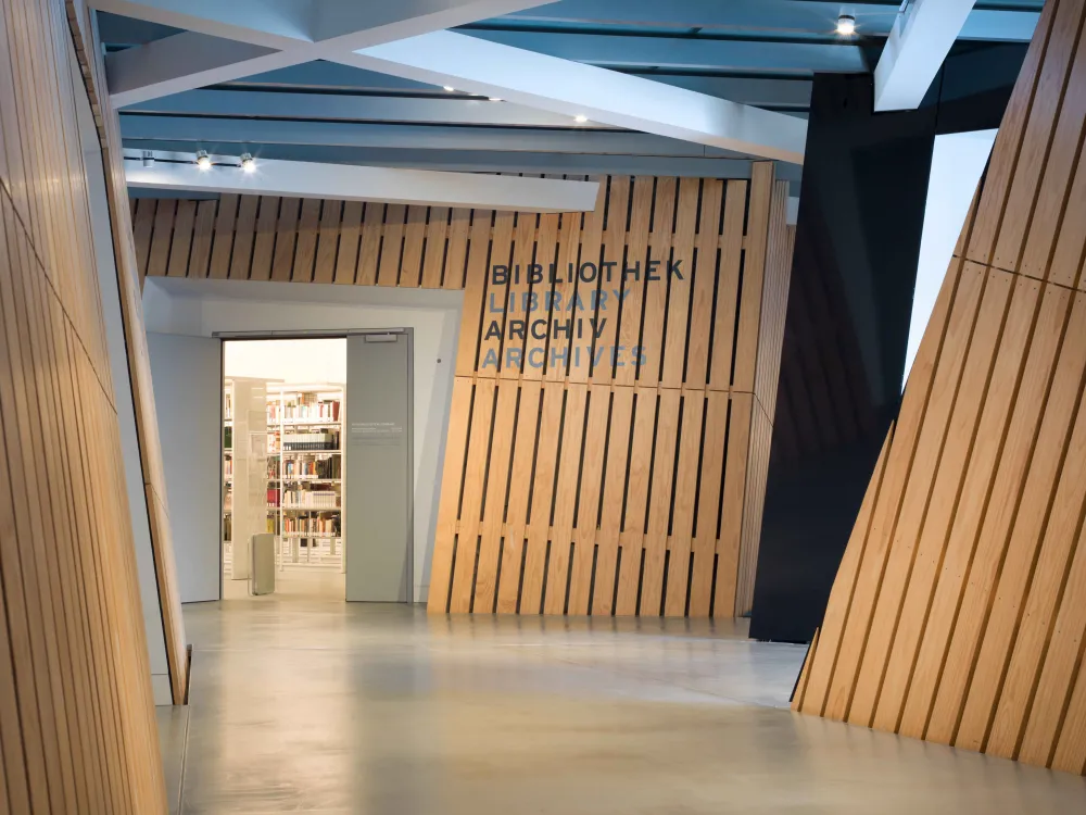Das Foto zeigt einen mit Holzpanelen verkleideten Flur, an dessen Ende sich eine geöffnete Tür befindet. An der Wand rechts neben der Türe steht "Bibliothek, Archiv".
