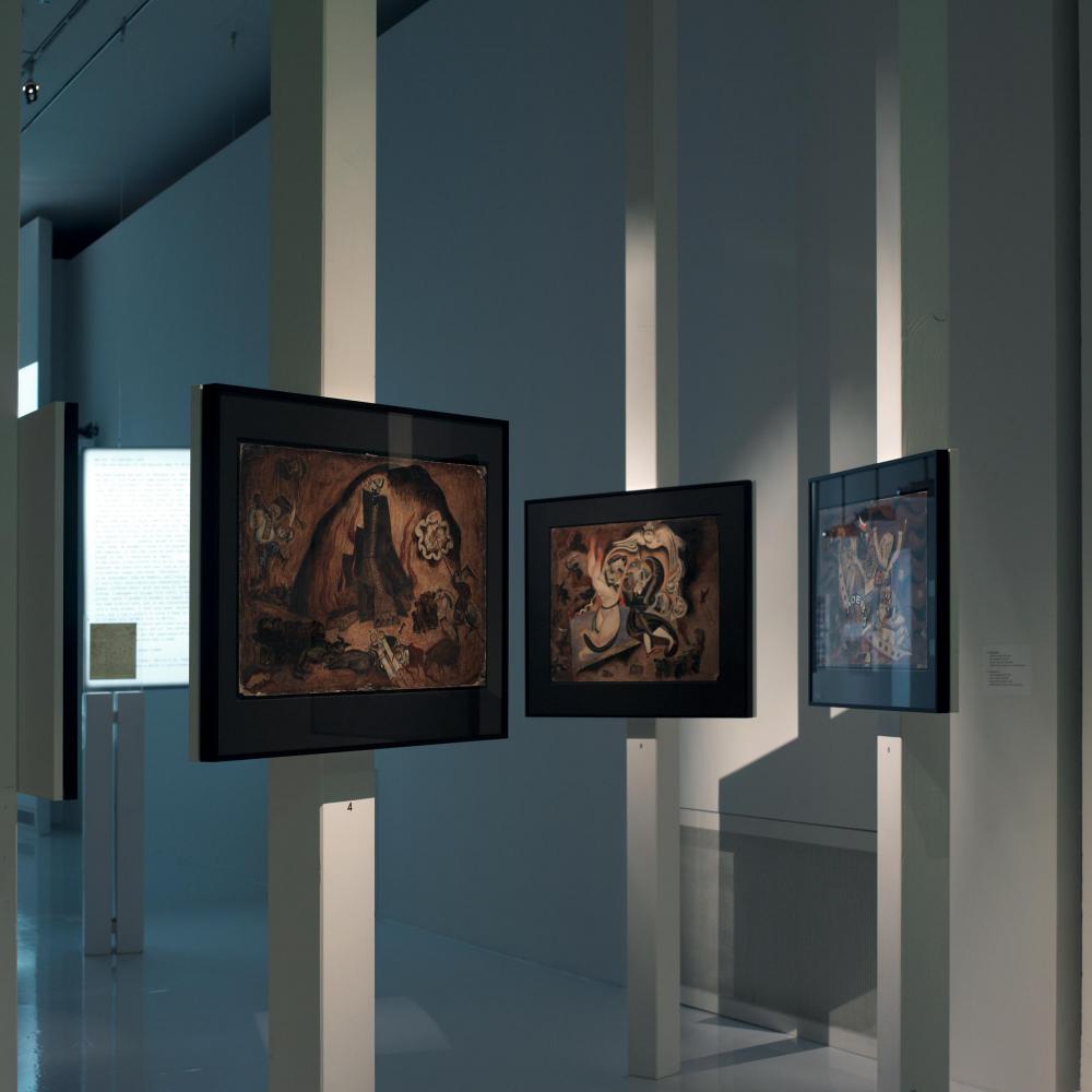 Mehrere Gemälde in einem Ausstellungsraum, die im Raum stehen und an Stelen befestigt sind