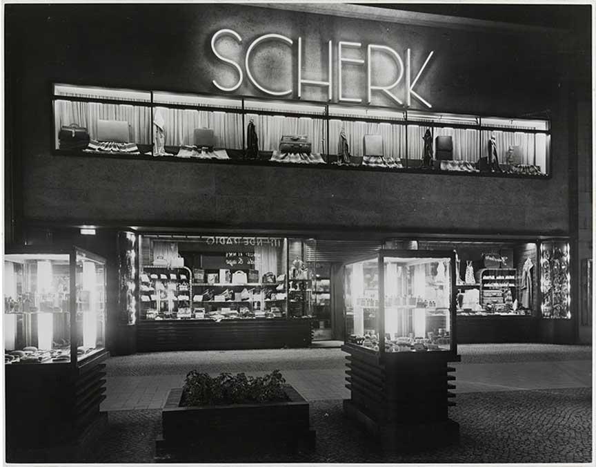 Black and white photo of a building with illuminated showcases and the glowing lettering Scherk on Kurfürstendamm at night.