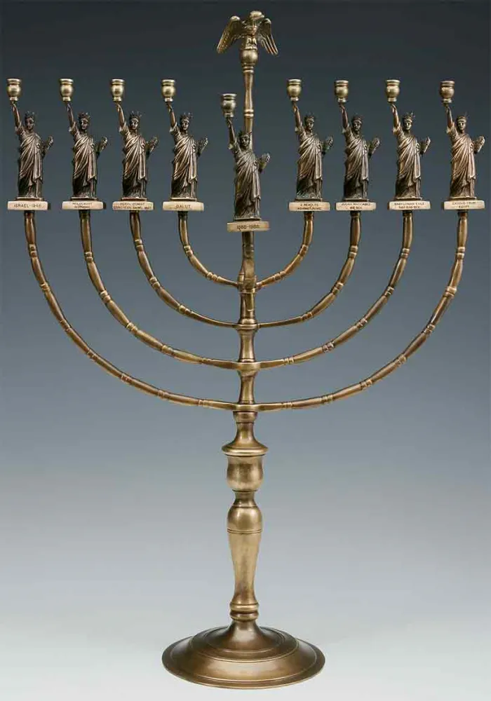 Leuchter mit runden Fuß, darauf ein aufmontierter Schaft, von dem rechts und links je vier Leuchterarme abgehen, die jeweils in einer Miniatur-Freiheitsstatue münden.