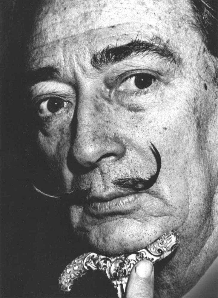 Close-up photo of Salvador Dali in black-and-white, his chin rests on the knob of a walking stick