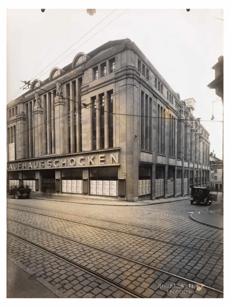 Eckansicht eines großen alten Gebäudes mit der großen Aufschrift „Kaufhaus Schocken“, im Vordergrund Straßenbahnschienen.