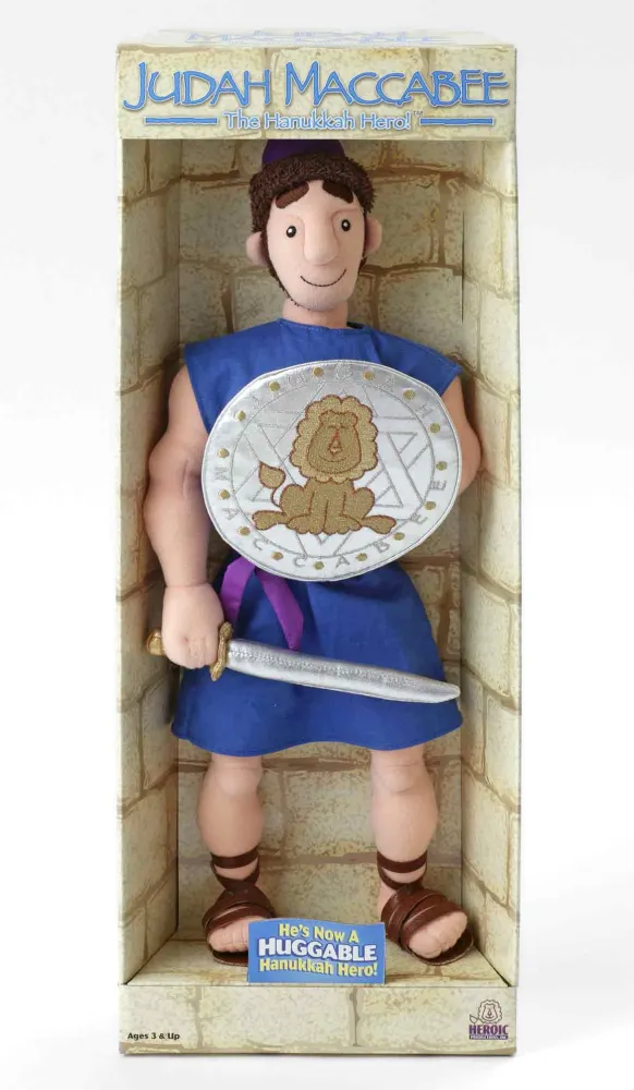 Judas Makkabäus-Stoffpuppe, noch im Verkaufskarton. Die Figur lächelt, ist mit einer ärmellosen, knielangen blauen Tunika und Römersandalen bekleidet, und trägt ein Schwert und einen Schild, der mit einem Löwen und einen Davidstern verziert ist.