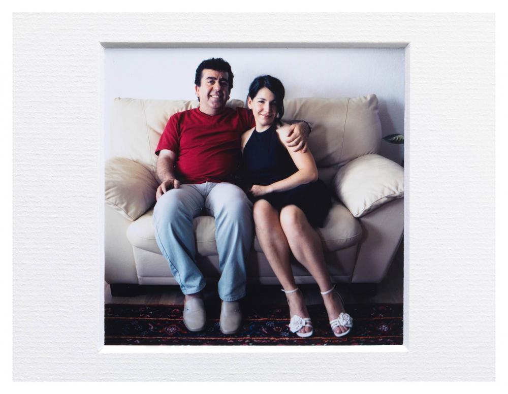 A woman and a man sit on a small couch and smile at the camera, the man has his arm around the woman's shoulders