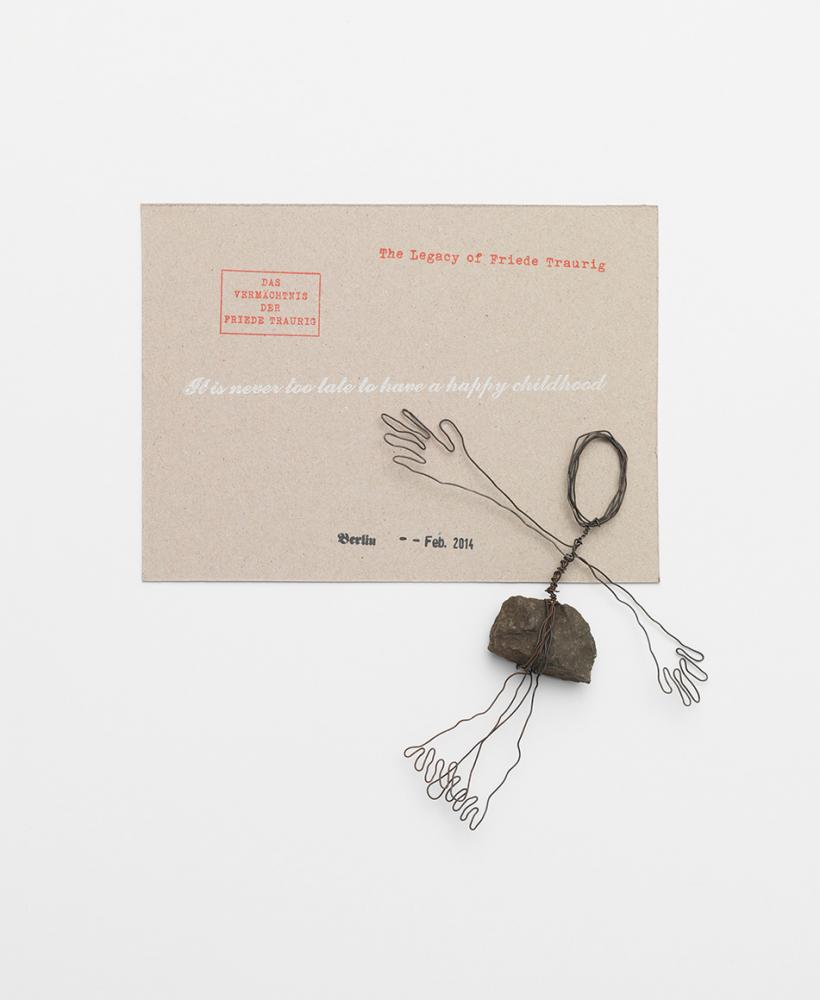 A small wire sculpture attached to a small stone depicting a human figure next to a tan card with text on it