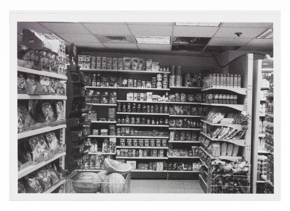 Black and white photograph of a grocery store aisle, a pile of watermelons is surrounded by boxes of food and snacks that line the shelves