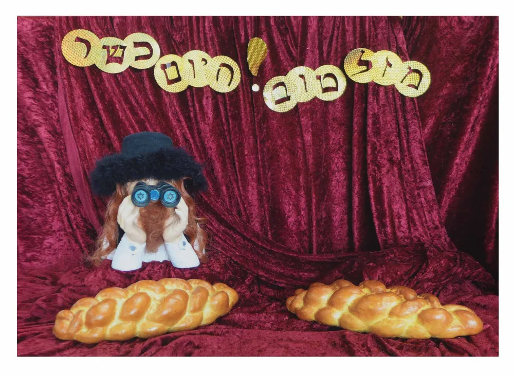 Golden paper Hebrew letters, a mannequin with binoculars, and two challah bread loafs are composed on red velvet fabric