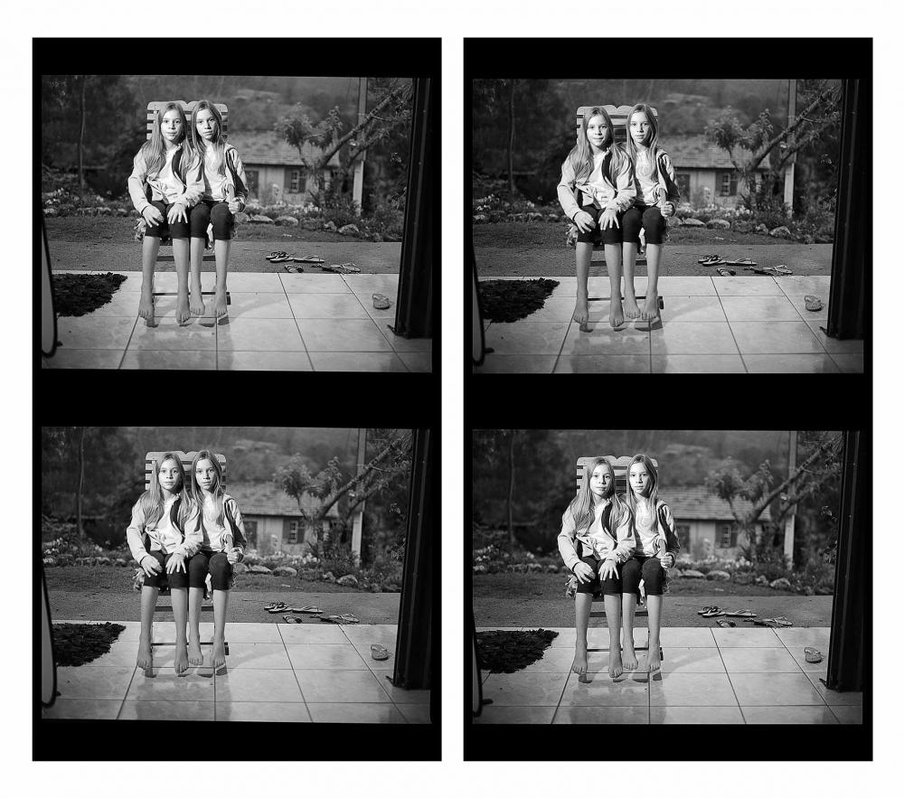 Eine vierteilige, sich wiederholende Schwarz-Weiß-Szene von zwei jungen Mädchen in identischer Kleidung, die nebeneinander auf demselben Holzstuhl sitzen.