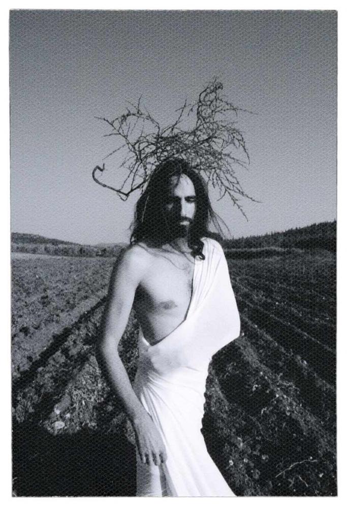 Photo: In the foreground, a man with long dark hair is standing in front of a fallow field with deep furrows and is wrapped tightly in a white cloth that leaves half his torso exposed. On his head, some foliage reminiscent of the crown of thorns.