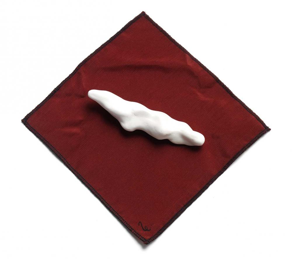 An oblong shape made of hard white plaster with a smooth surface and irregular round protrusions, possibly a worry stone due to its size and texture, on a square of wine-red fabric