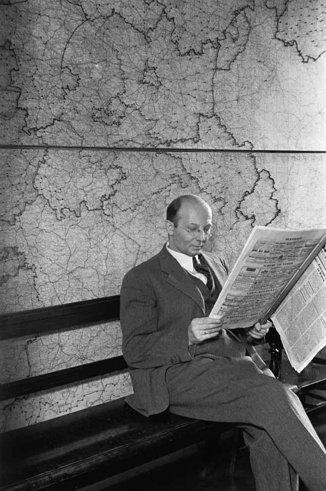 Black and white photography: a seated man reading an issue of the C.V. newspaper. There is a map on the wall behind him.