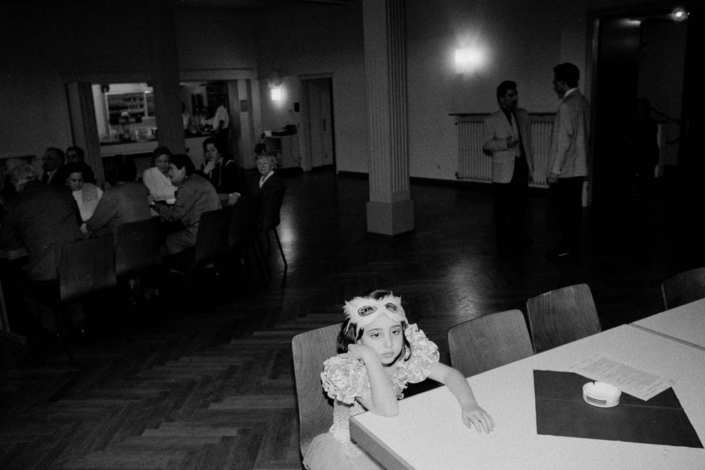 A black and while photo of a young girl wearing a frilly, puffy dress and a feather eye mask on her forehead, she is sitting at a table by herself with her head in her hand