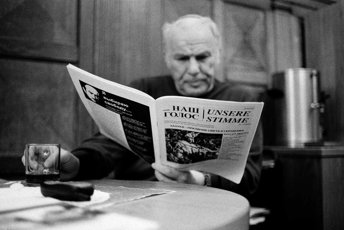 A black and white photograph of a man sitting at a table with a newspaper in his left hand and a cup in the other