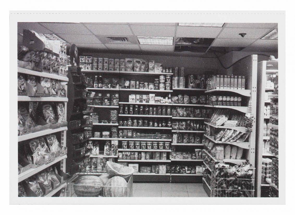 Black and white photograph of a grocery store aisle, a pile of watermelons is surrounded by boxes of food and snacks that line the shelves 