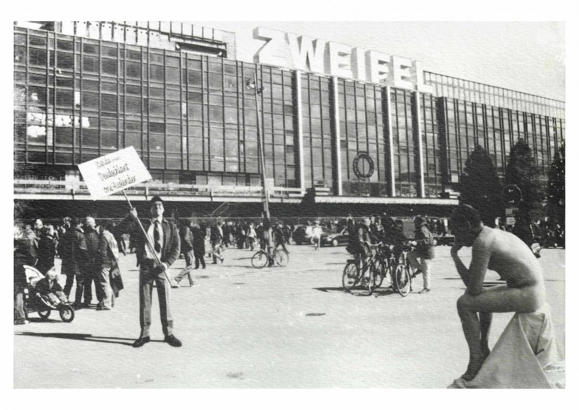 Eine historische Schwarz-Weiß-Fotografie einer Menschenmenge vor einem großen Gebäude. Rechts ist ein nackter Mann zu sehen, der auf einem Tuch sitzt, links hält ein Mann ein Mahnwachenschild.