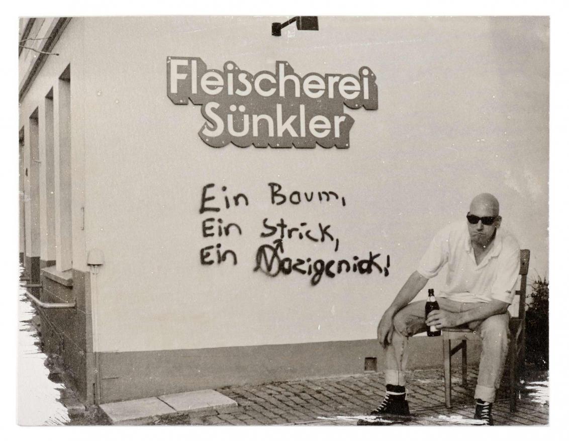 Ein Mann mit Sonnenbrille und einem Bier in der Hand sitzt neben einer Wand mit Graffiti.