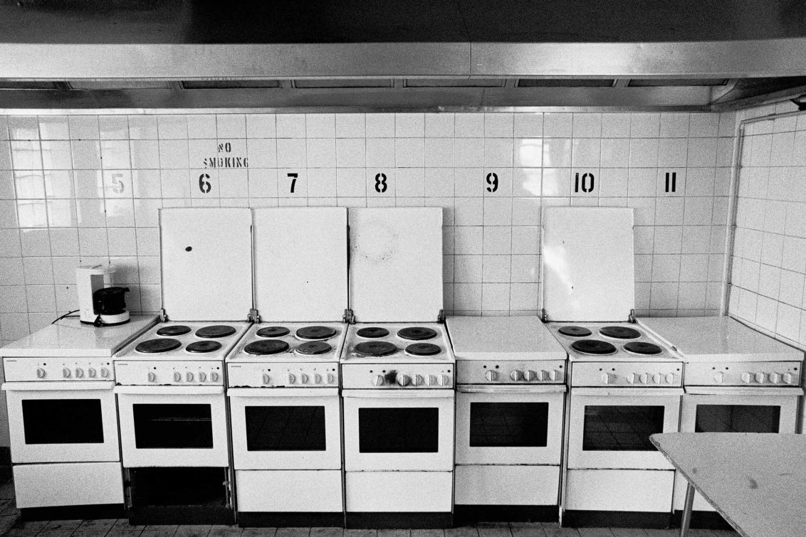 A black and white photograph of a row of small white ovens, some of the ovens have their stovetop open, all the ovens have a black number on the wall above them next to a “No Smoking” sign