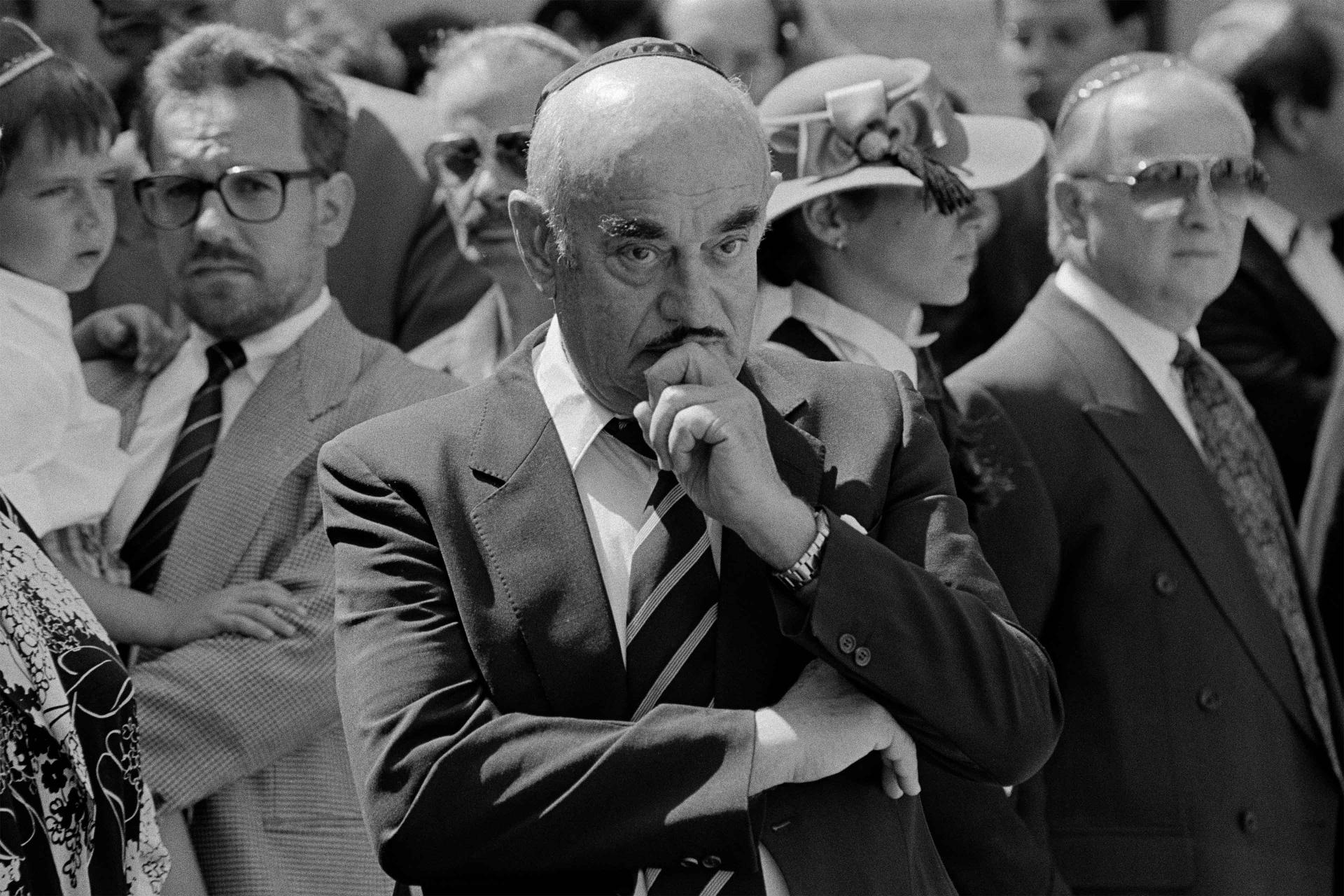 Black-and-white photograph: Artur Brauner stands among other mourners, holding his left hand to his chin and angling his right arm