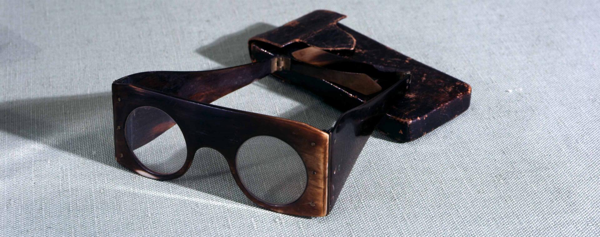 Quadratische Brille mit runden Gläsern, daneben ein passendes Etui.