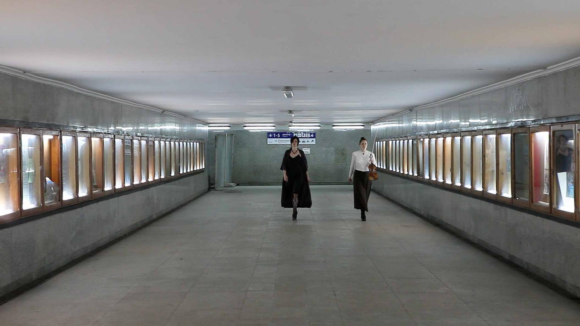 Two woman, one holding a violin and bow, walk towards the camera in an empty concrete hallway
