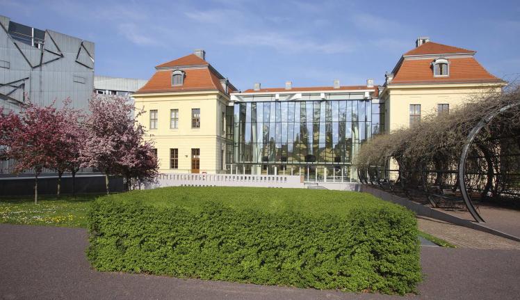Altbau und Glashof des Jüdischen Museums Berlin vom Garten aus, rechts Laubengang, links blühende Bäume.