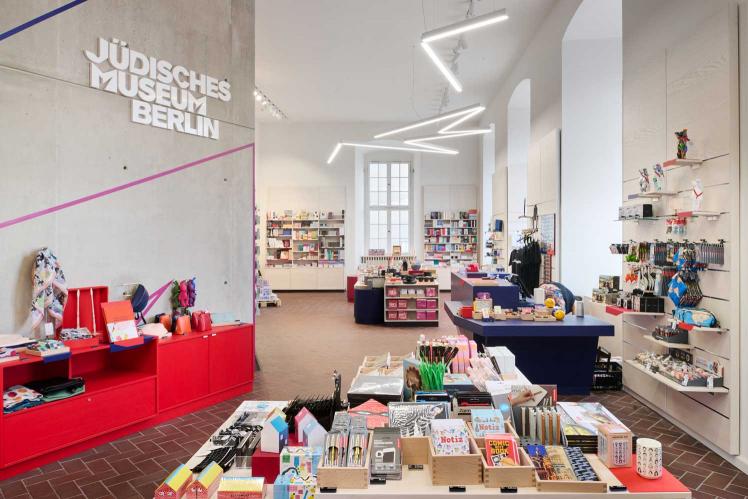 Shop area with brightly colored furniture and a colorful assortment, the white logo with the lettering Jewish Museum Berlin can be seen on a concrete wall, and a neon lamp on the ceiling that zigzags through the room.