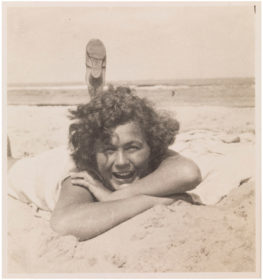 Black and white photo of a curly woman on the beach lying on her belly in the sand with a foot stretched out into the sky.