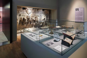 A look into an exhibition room. On the rear wall, a plaster cast of the Arch of Titus can be seen. In the foreground stand a glass case with a menorah, Torah rimonim, and open books.