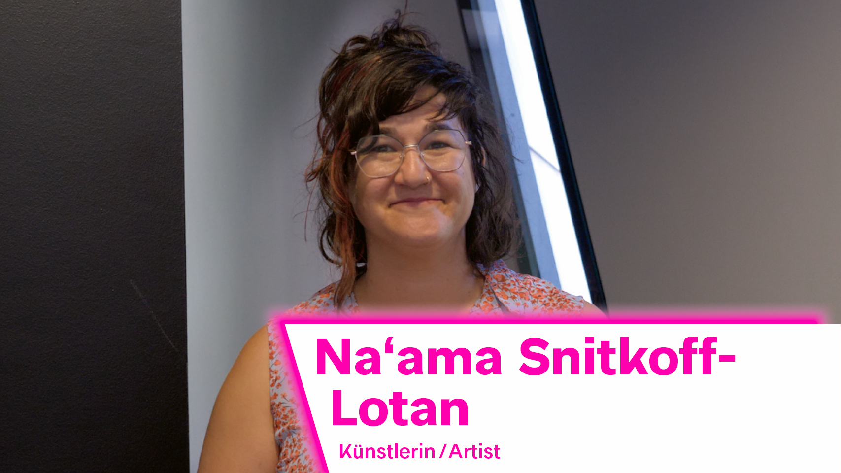 The image shows Israeli artist Na'ama Snitkoff-Lotan. She is wearing a blouse with a colourful design, her brown hair is loosely held together at the back of her head and she is waeting glasses.