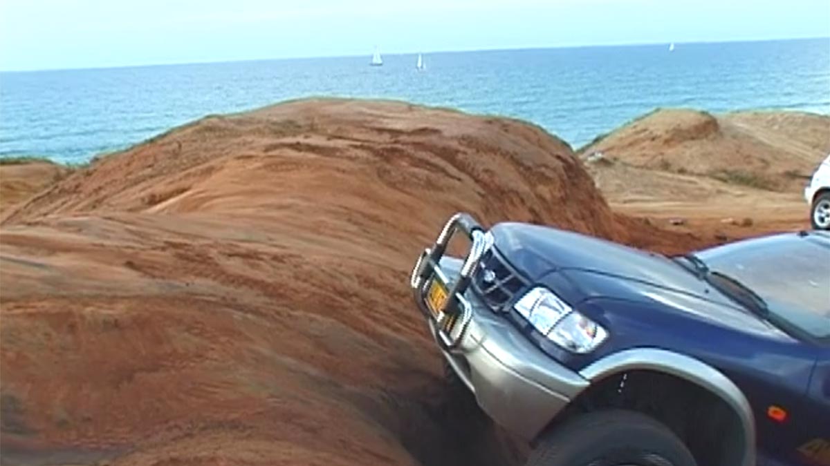 A blue off-road vehicle drives up a sand dune, the sea can be seen in the background.