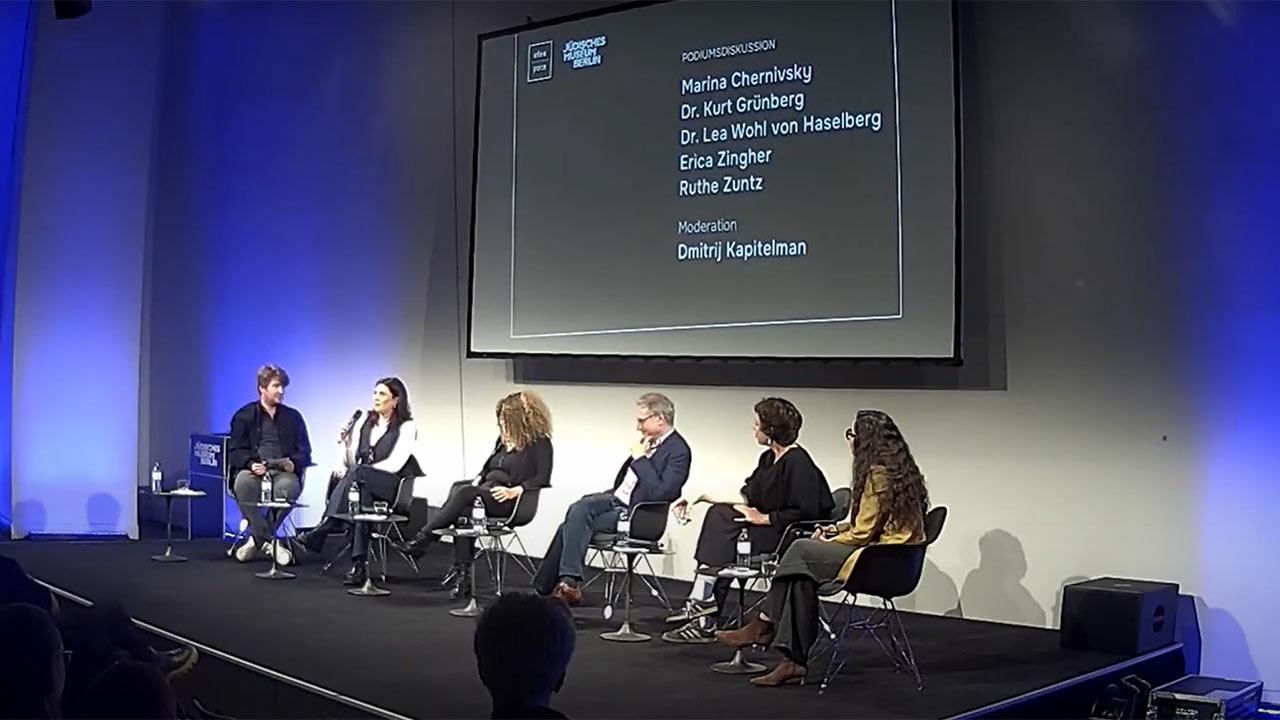 Six people sit on chairs on a stage, one person holds a microphone; in the background is a screen listing the panelists.