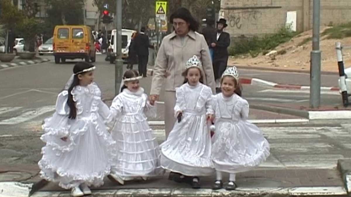 A woman leads four young girls in white wedding dresses across a street.