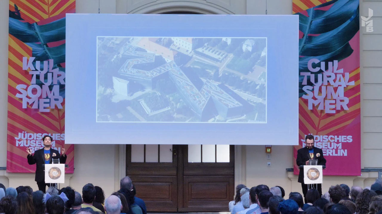 Stage in the glass courtyard of the JMB with flags from the Cultural Summer, a screen with a picture of the Libeskind Building and a person at a lectern to the left and another person on the right of the screen.