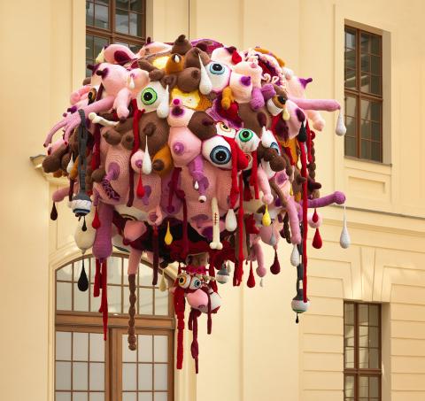 A large ball of crocheted body parts hangs from the ceiling of the glass courtyard. The ball consists of pink, purple, brown and white crocheted eyes, penises, breasts and bones.