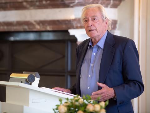 A man with gray hair, jacket and shirt, stands at a lectern and gives a speech. 