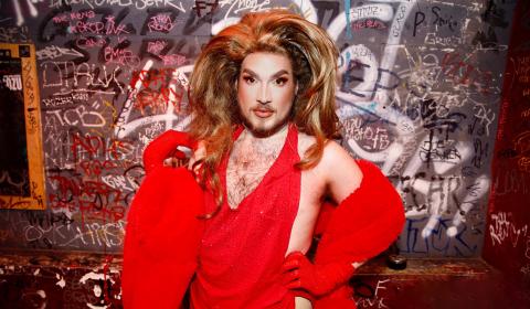 Drag DJane Jewels, in a red dress, poses in front of a graffiti wall.