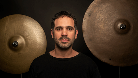 A man in a black sweater stands in front of a wall with drum cymbals hanging on it.