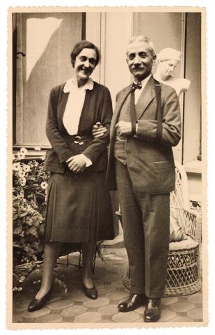 Black and white photo of two people in the entryway of a house with flowers, a statue, and a wicker chair in the background 